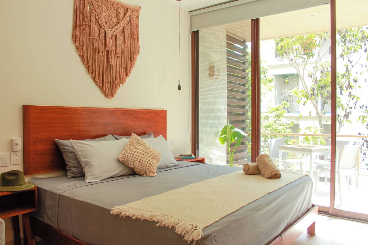 A bedroom with a wooden headboard, a bed with grey bedding, a macrame wall hanging, and a view of an outdoor area with a table and chairs.