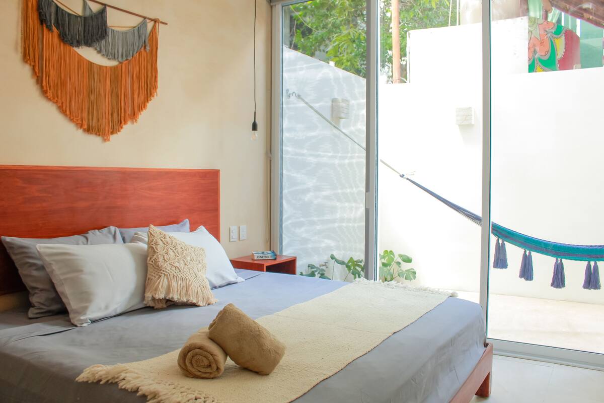 A bedroom with a bed, pillows, a blanket, a macrame wall hanging, and a view through a glass door.