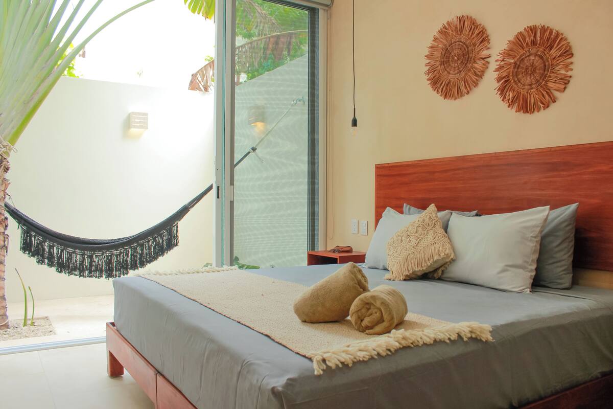A bedroom with a bed, pillows, a bedside table, a hammock, and a view of the outdoors.