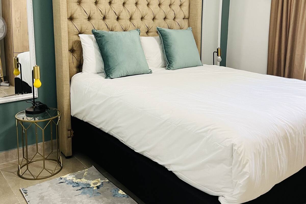 A neatly made bed with a tufted headboard, a bedside table with a lamp, and a patterned rug on a tiled floor.