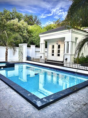 Una piscina rectangular con borde negro, rodeada por una cerca blanca y un edificio blanco con columnas y ventanas.