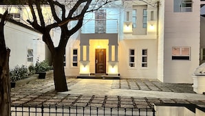 A modern two-story house with a well-lit entrance, a large tree in the foreground, and a small outdoor seating area with a table and bench.
