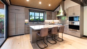 A modern kitchen with a large island, bar stools, a stainless steel range hood, and a window with a view of greenery. - Luxury Nohea Home w/ Pool & private 2 BR Ohana
Access to Mauna Lani Sports Club! (Kamuela)
