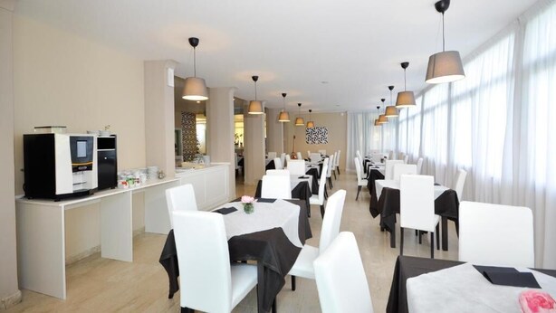 En restaurant i et hotel med hvide borde og stole, hængende lys og en køkkenbar med hårde hvidevarer.