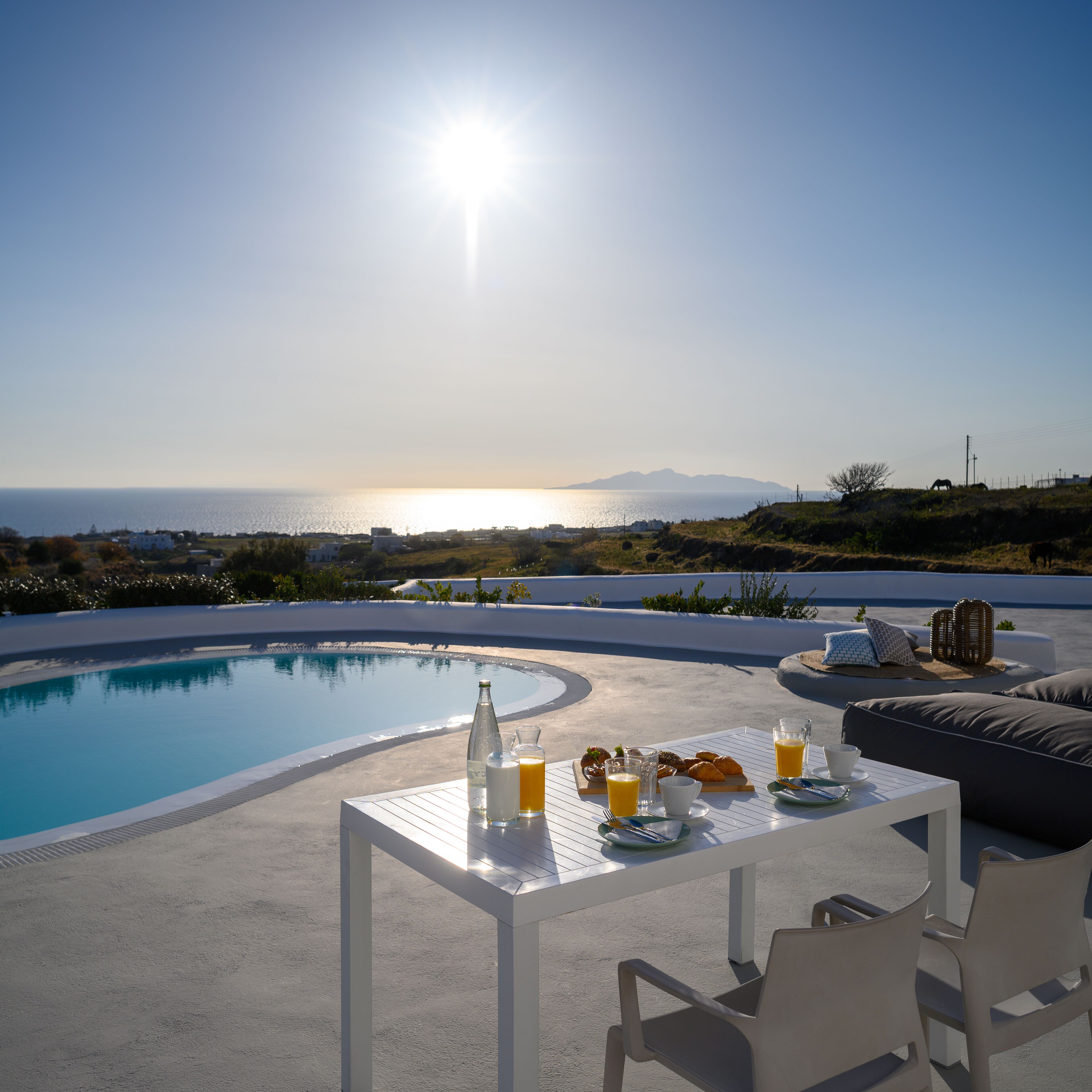 Zona de comedor junto a la piscina con una mesa preparada para el desayuno, con vista a un cuerpo de agua y un paisaje montañoso.