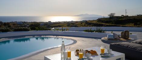 Zona de comedor junto a la piscina con una mesa preparada para el desayuno, con vista a un cuerpo de agua y un paisaje montañoso.