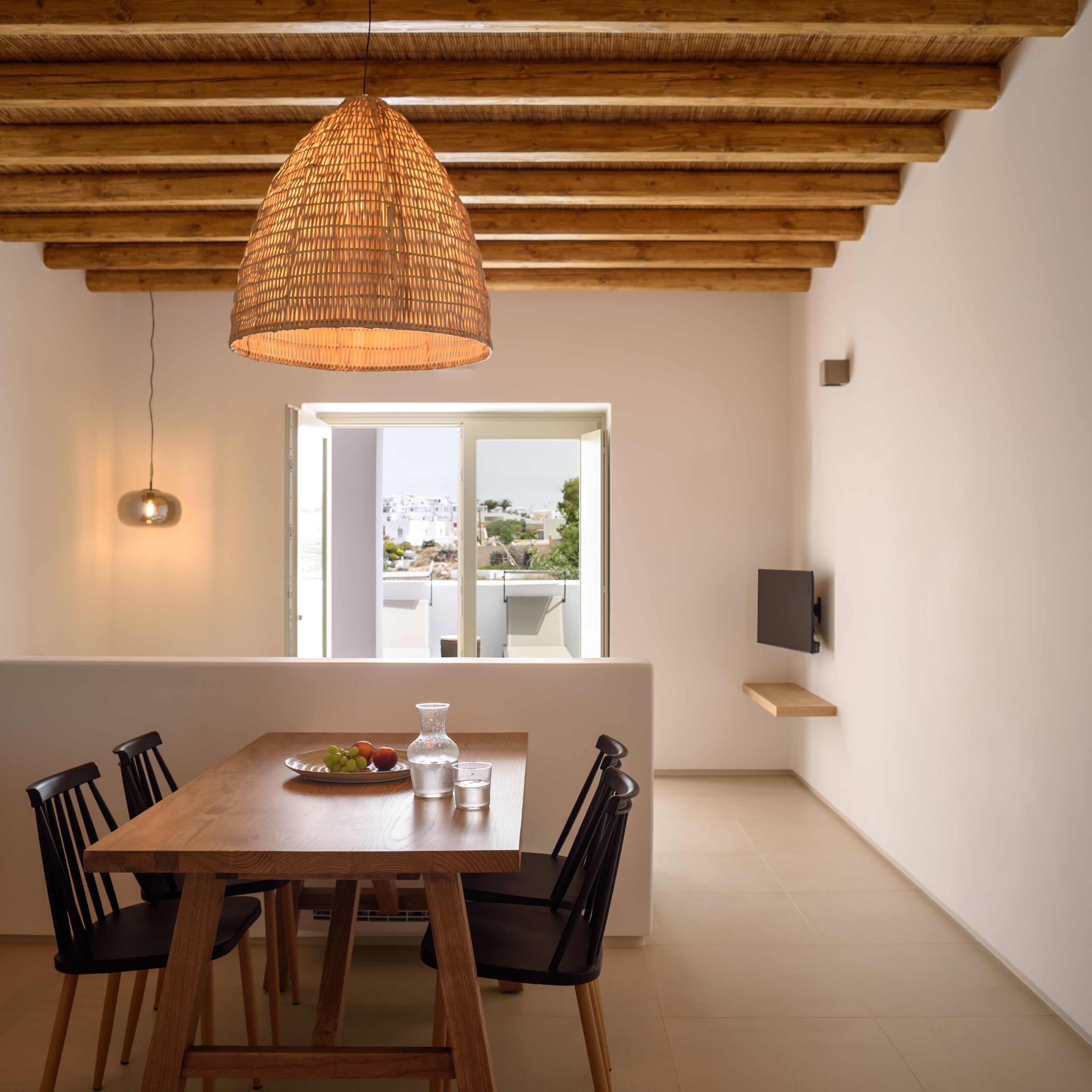 Uma sala de jantar com mesa de madeira, cadeiras, luminária pendente e vista para o exterior através de uma grande janela.
