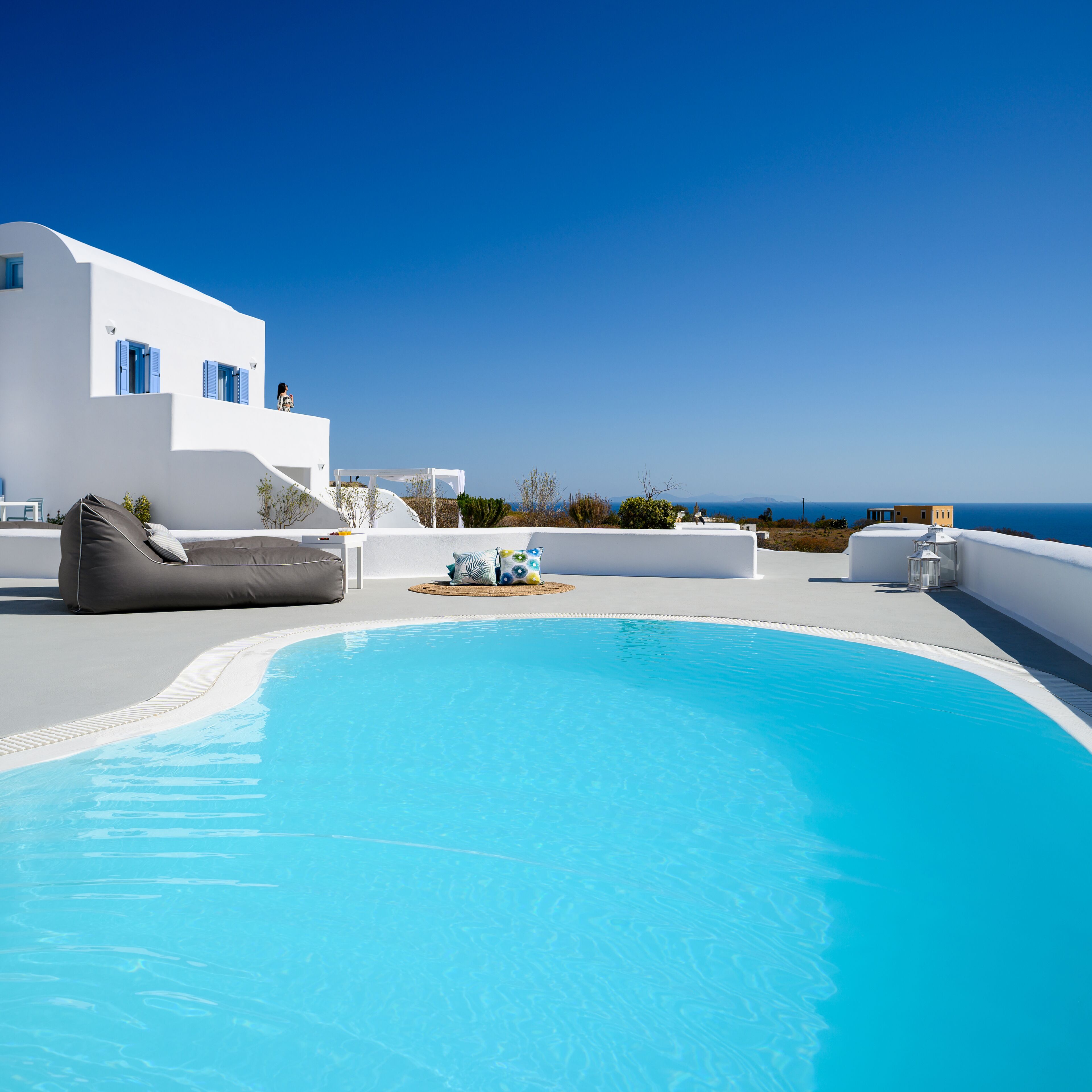 A pool area with a white building, a lounge chair, and a view of the sea.