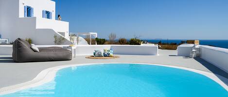 A pool area with a white building, a lounge chair, and a view of the sea.