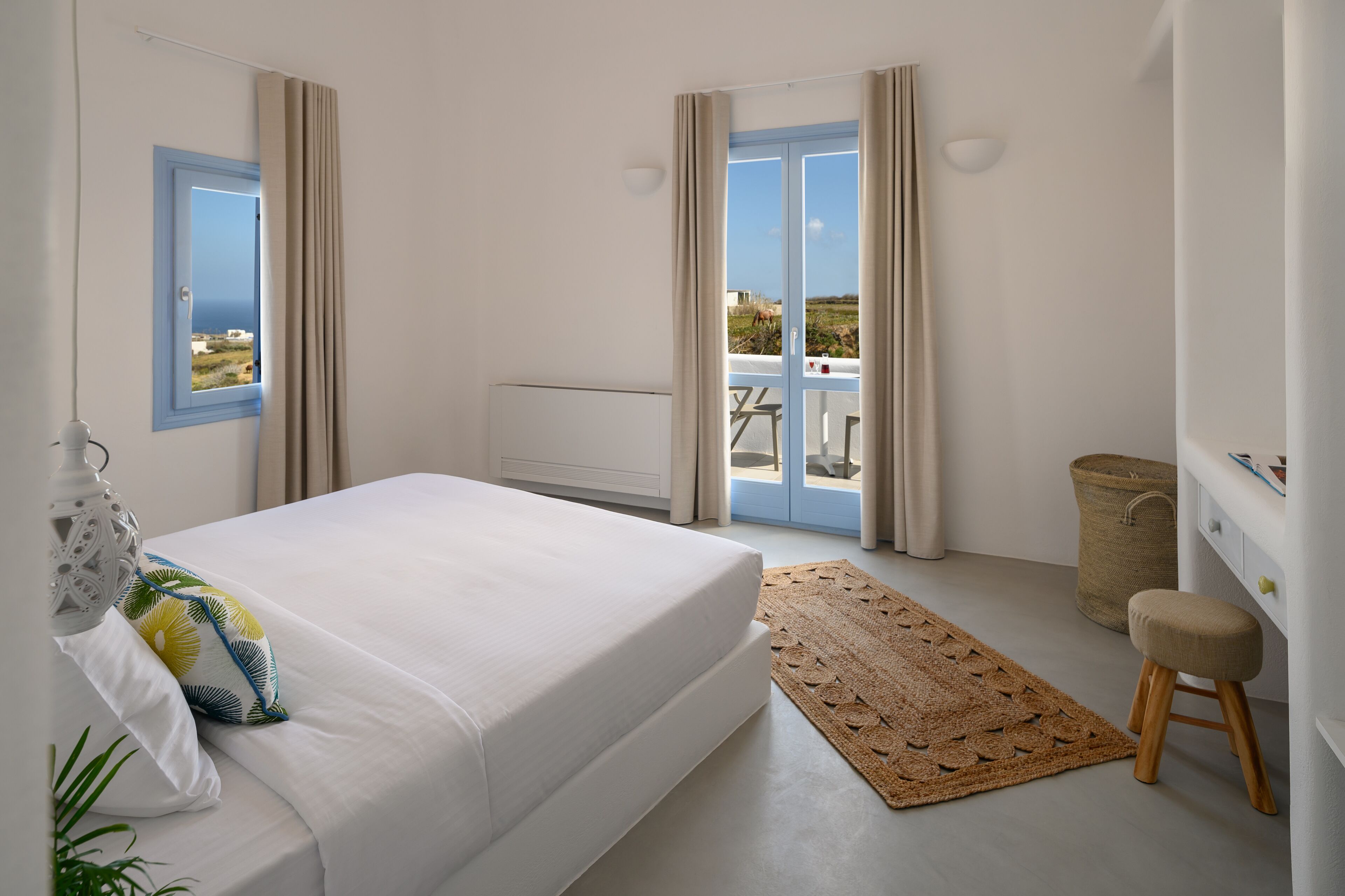 A bedroom with a bed, a small stool, a waste basket, and a view of the outdoors through a door.