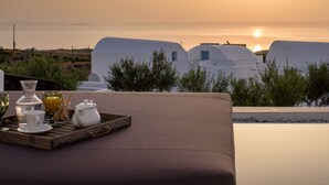 A terrace with a table set for a meal, overlooking a coastal village with white buildings and a sunset. - Este Luxury Suites (Santorini)