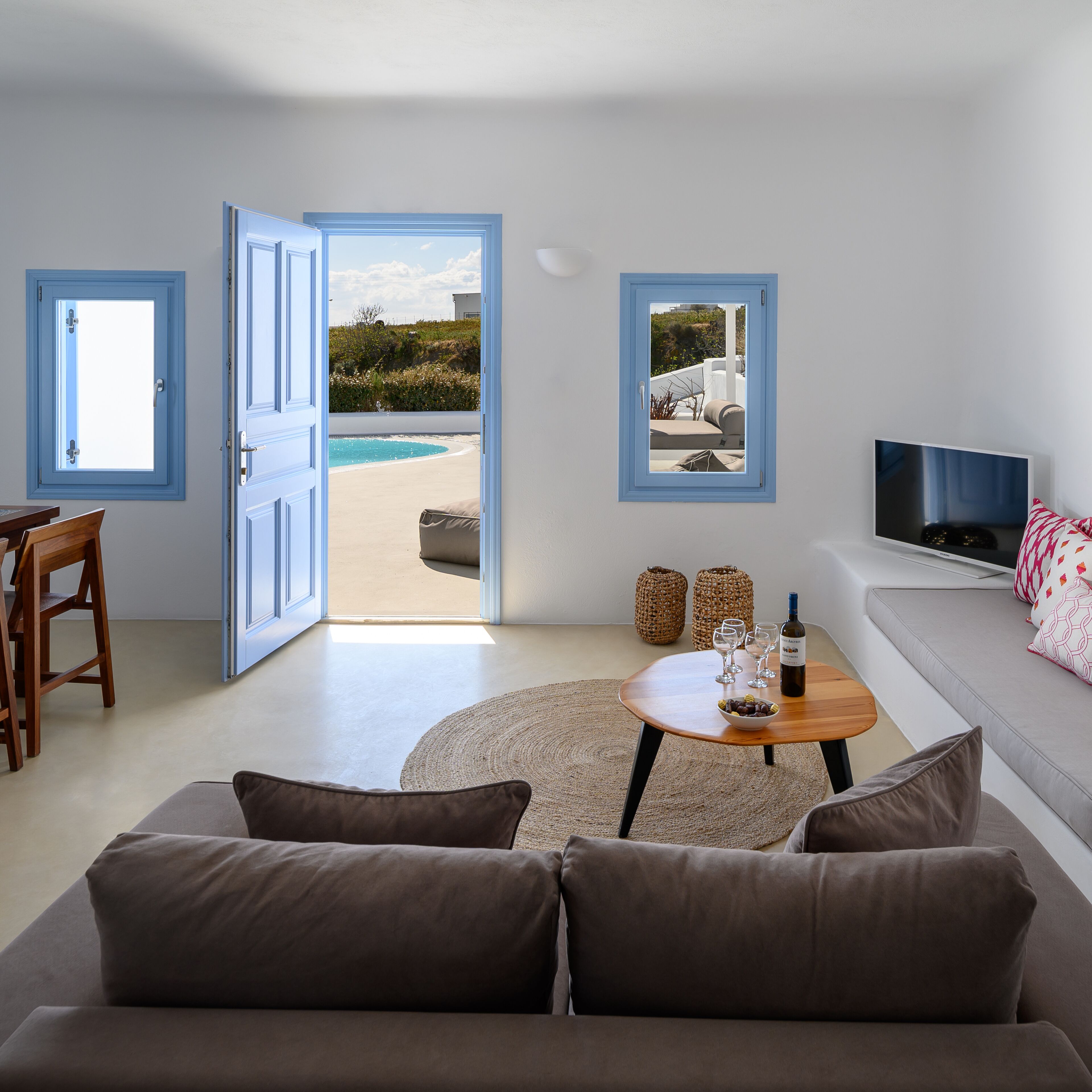 A living room with a sofa, a coffee table with wine glasses and a bottle, a TV, and two doors leading to a pool area.