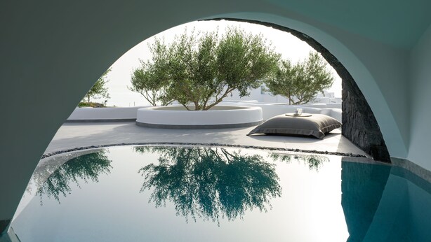 A clear view of a swimming pool with a reflection of trees and a white building in the background. - Este Luxury Suites (Santorini)