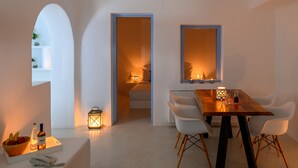 A modern dining area with a wooden table, white chairs, and a small side table with a bottle and a bowl of fruit. - Este Luxury Suites (Santorini)