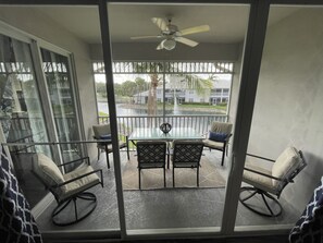 Een balkon met een glazen tafel en stoelen, een plafondventilator en uitzicht op een waterpartij en gebouwen.