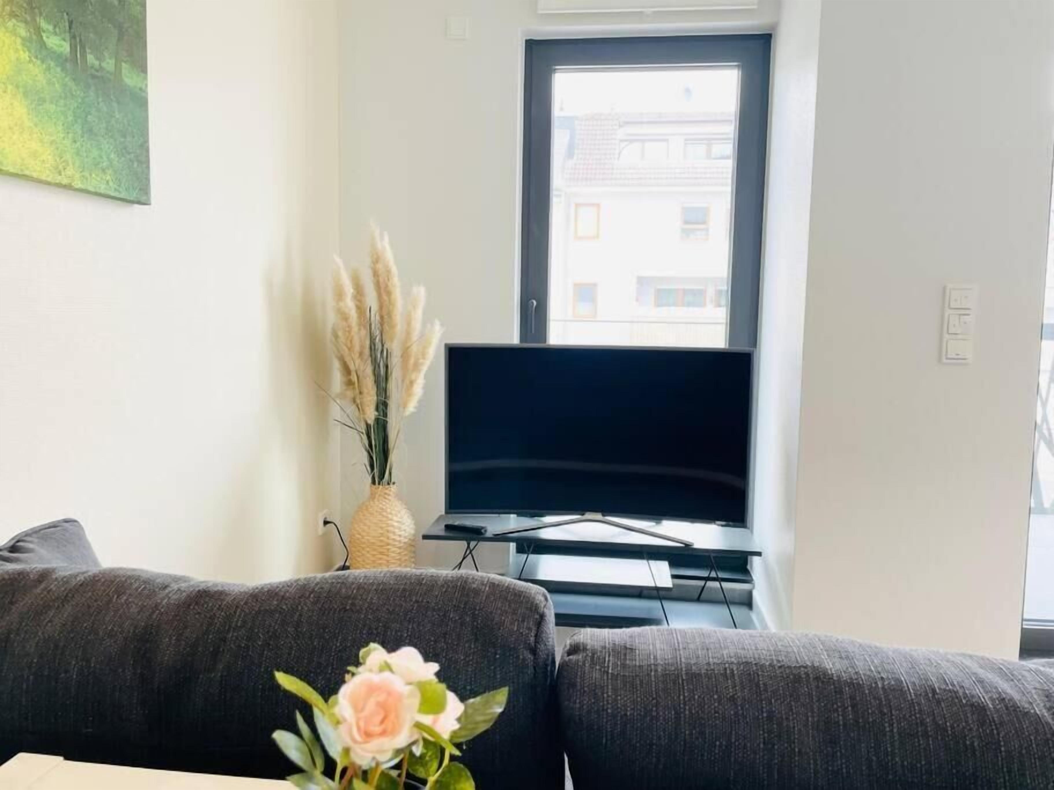 Una sala de estar con un televisor de pantalla plana, un sofá gris, una mesita y un jarrón con pastos secos y una flor rosa.