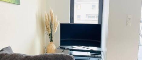 Una sala de estar con un televisor de pantalla plana, un sofá gris, una mesita y un jarrón con pastos secos y una flor rosa.
