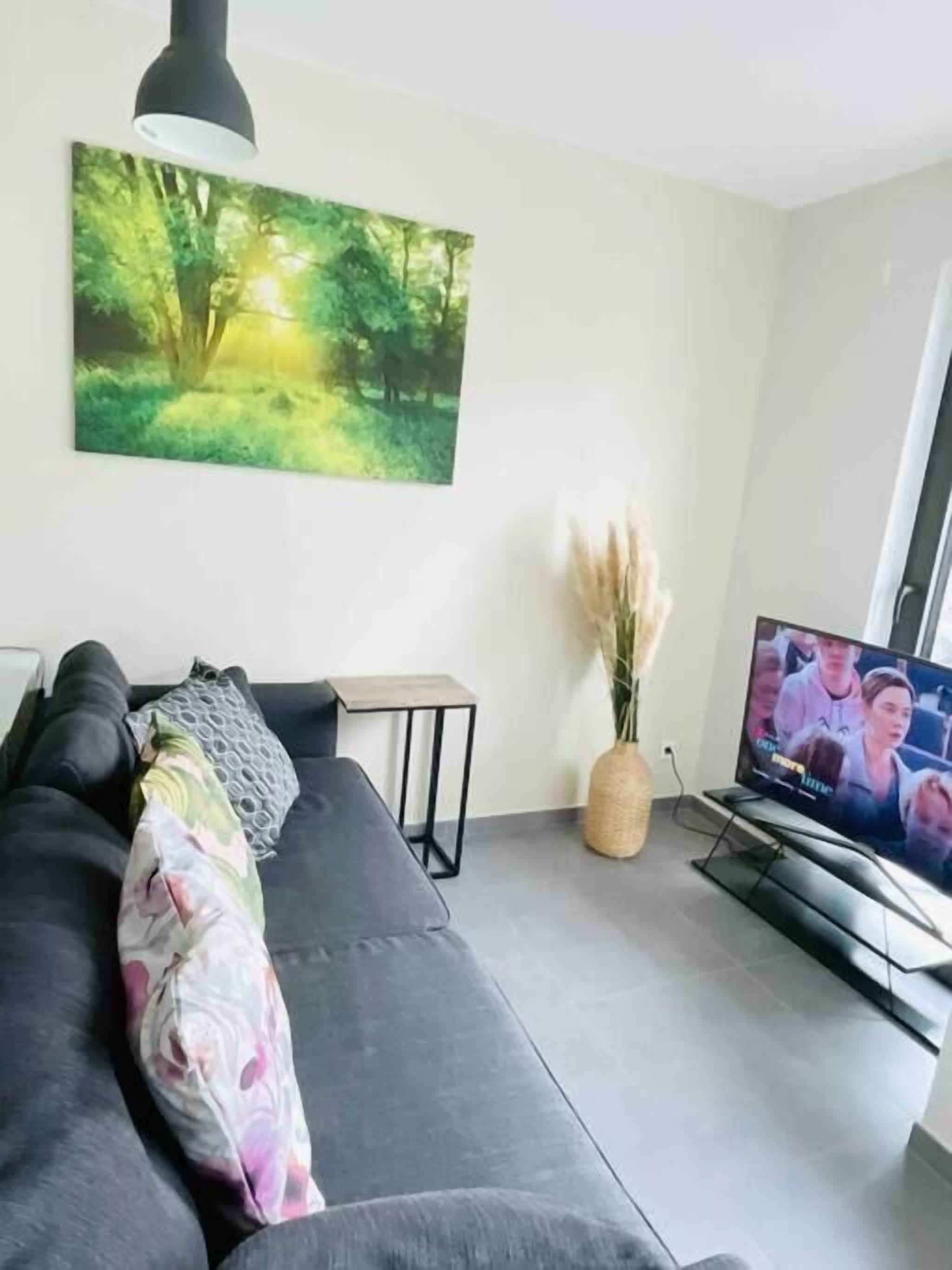 A modern living room with a dark sofa, a painting of a forest, a small table, a vase with pampas grass, and a TV displaying a show.