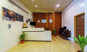 Un área de recepción con un mostrador de madera, un televisor de pantalla plana, un ventilador de pared y diplomas enmarcados. Hay plantas en macetas y una puerta de madera con un número.