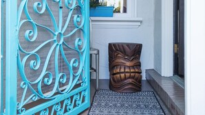 Une porte en métal turquoise, une porte en bois, une fenêtre agrémentée d’un jardinier bleu et un tapis à motifs posé sur un sol en briques.