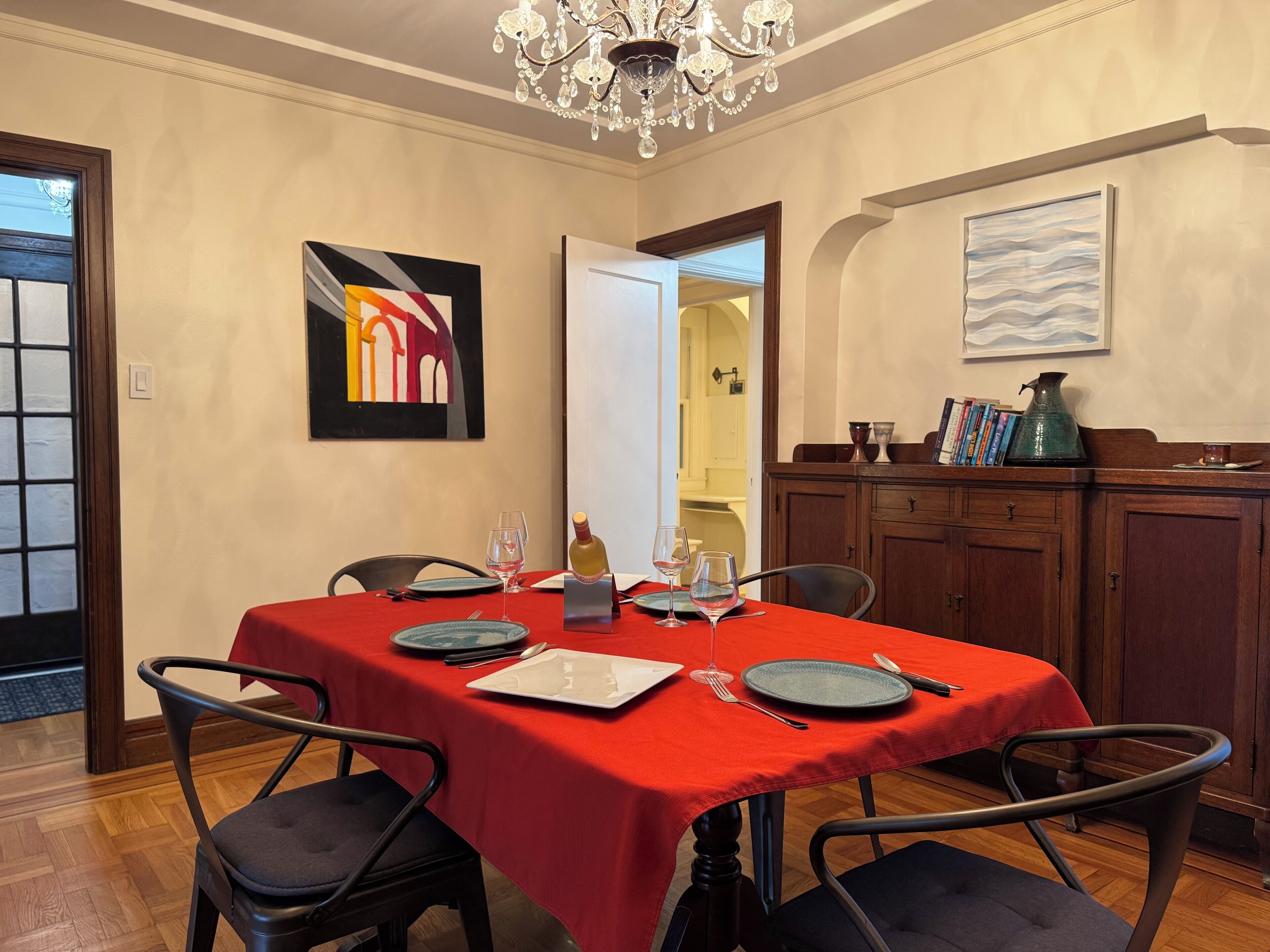 A dining room with a red tablecloth, wooden chairs, a chandelier, and artwork on the walls.