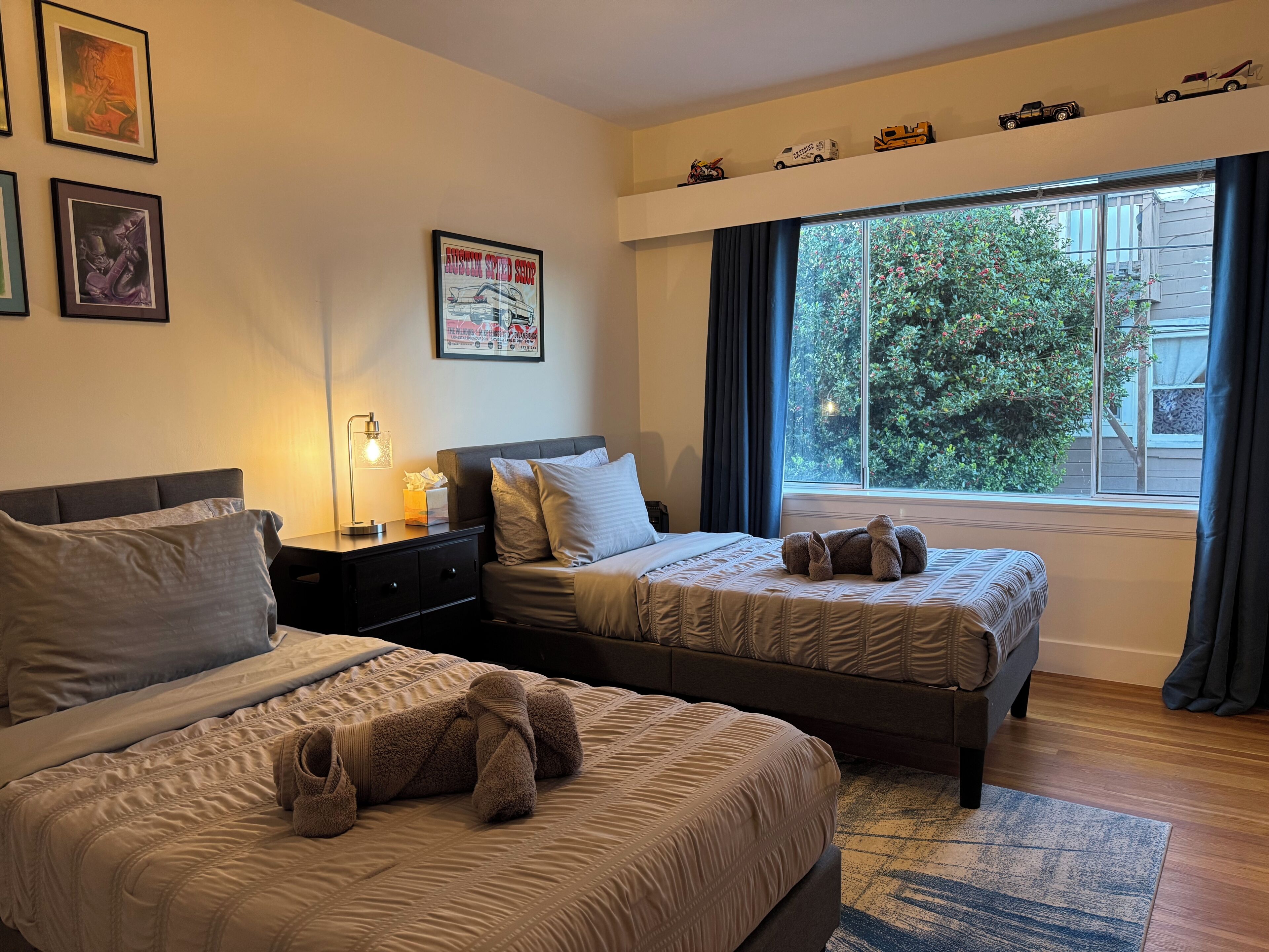 Een hotelkamer met twee bedden, een nachtkastje met een lamp, een poster aan de muur en een raam met uitzicht op bomen.