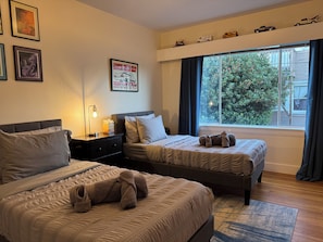 Habitación de hotel con dos camas, una mesita de noche con lámpara, un cartel en la pared y ventana con vistas a árboles.