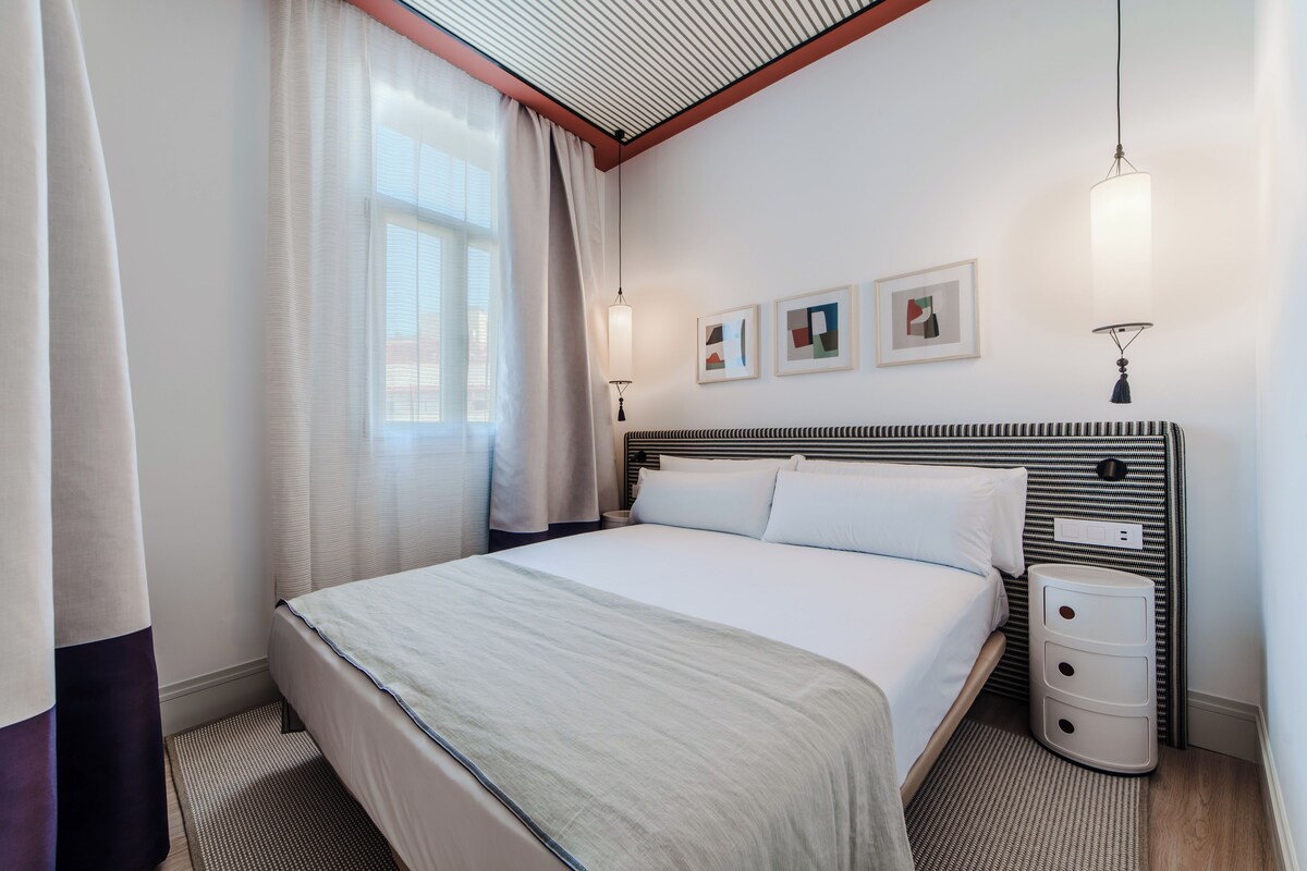 Une chambre d’hôtel bien rangée, avec un grand lit, une table de chevet, une commode et une lampe fixée au mur.