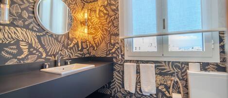 A bathroom with a black and gold leaf-patterned wall, a round mirror, a dark countertop, a white sink, a white toilet, and a window with a view of buildings.