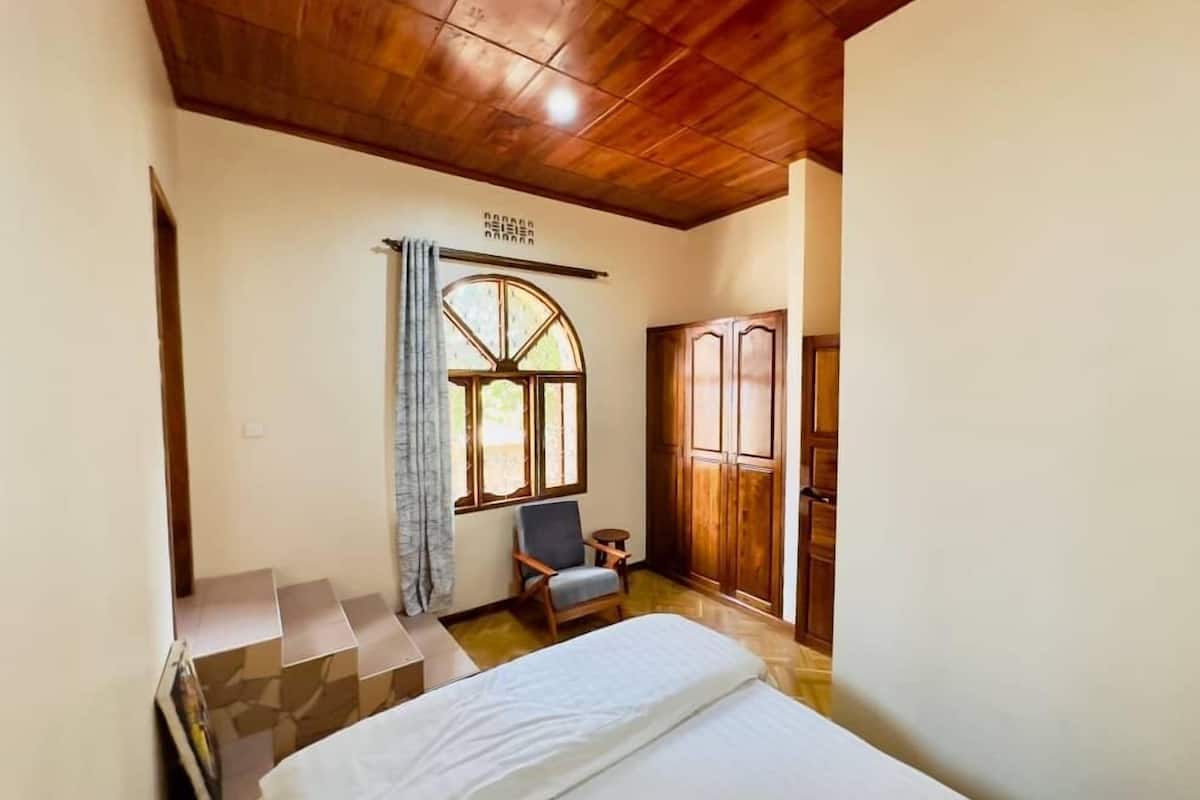 A bedroom with a wooden ceiling, a bed, a chair, a window with curtains, and a wardrobe.