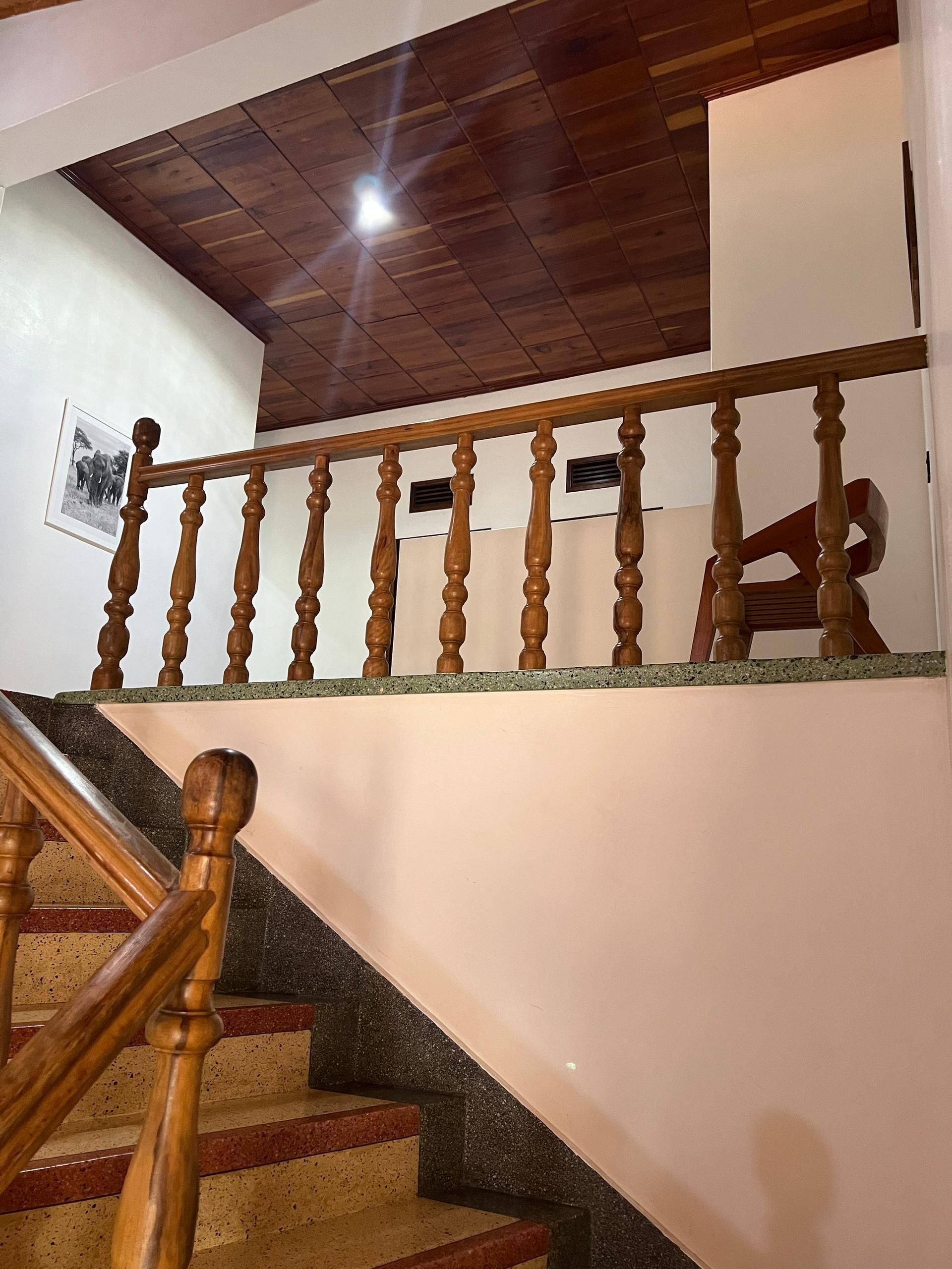 Un escalier en bois avec une rampe, un plafond avec des poutres apparentes et un mur orné d’un tableau encadré.