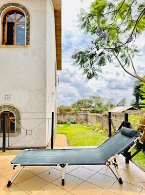 A sunbed in a covered outdoor area with a view of a garden and a building with arched windows. - Starehe Villa (Arusha)