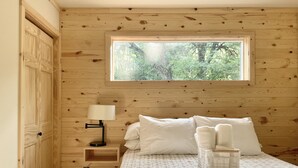 A bedroom with a wooden wall, a window with a view of trees, a bed with white bedding, and a bedside table with a lamp. - Modern Design - Fire Pit - Lake Access (Montello)