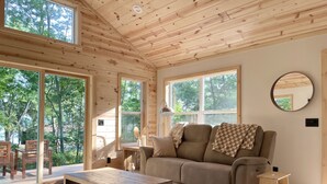 A cozy cabin interior with a wooden ceiling, large windows, a sofa, a coffee table, and a small wooden stool. - Modern Design - Fire Pit - Lake Access (Montello)