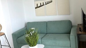 Ein Wohnzimmer mit einem grünen Sofa, einem weißen Couchtisch mit einer Pflanze, Holzboden und einem Strandbild an der Wand.