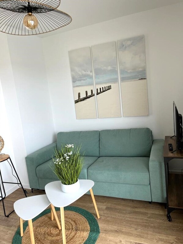 En stue med en grøn sofa, et hvidt sofabord med en plante, trægulv og et maleri af en strand på væggen.