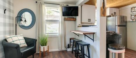 A compact living space with a sofa, a TV, a small kitchen area, and a trash bin.