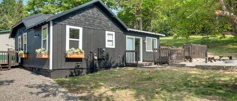 A small, dark-colored cabin with a sloped roof, a front porch, and a fenced yard.
