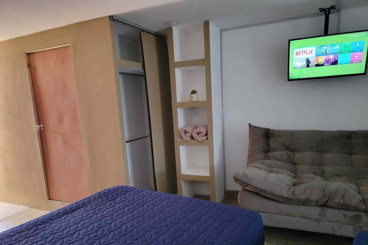 Quarto de hotel com cama, sofá, televisão fixada na parede e porta de madeira.