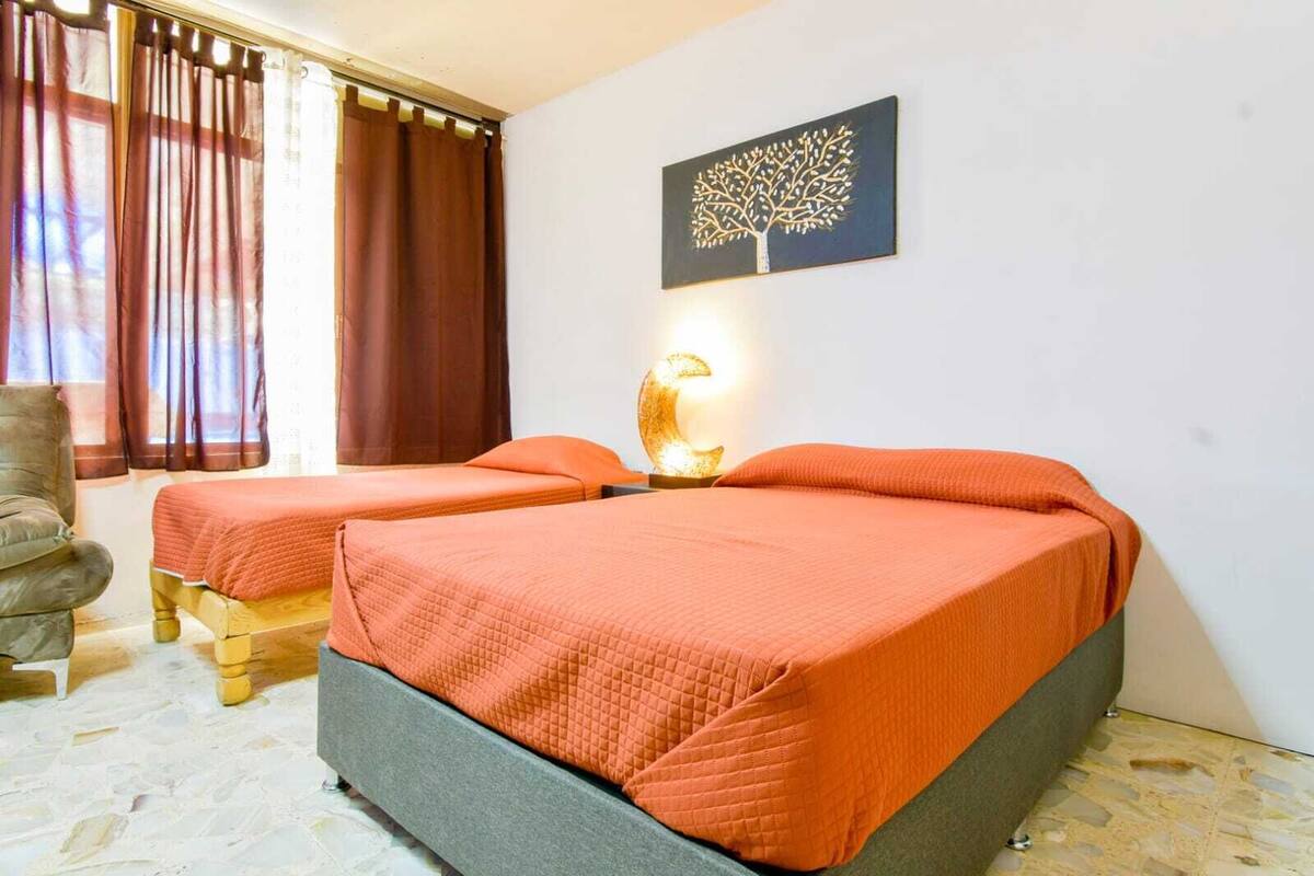 Quarto com duas camas, uma cadeira, uma luminária na parede e um quadro de uma árvore.