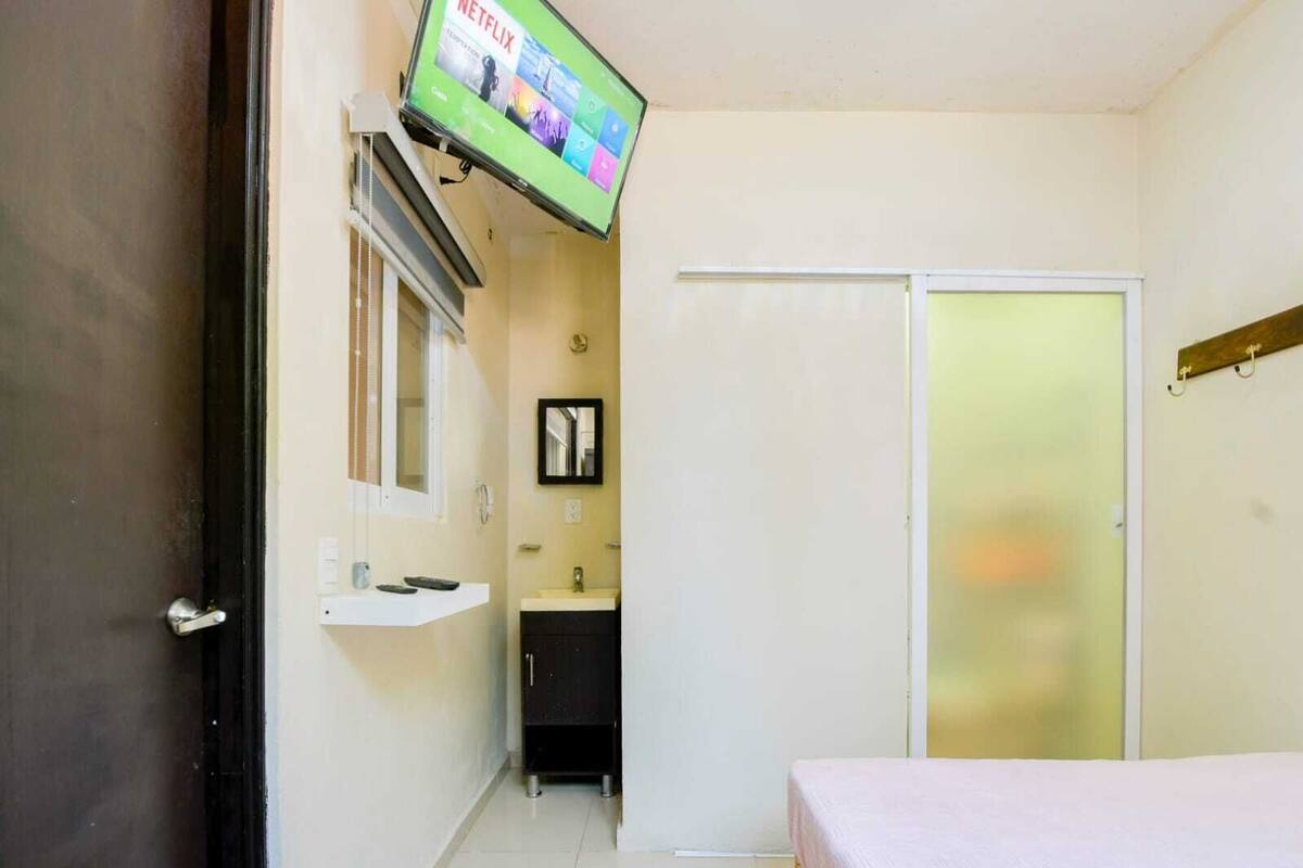 Quarto pequeno e compacto com uma cama, televisão fixada na parede, banheiro com pia e espelho e um armário.