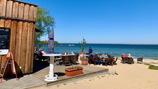 A wooden building with outdoor seating by a lake, tables, chairs, and a signboard. - Cozy vacation home with sauna, fireplace, garden and boat (pet-friendly) (Röbel/Müritz)