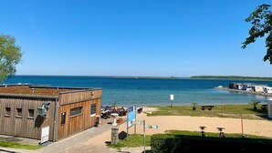 A coastal view with a wooden building, a sandy beach, and a clear blue sky. - Cozy vacation home with sauna, fireplace, garden and boat (pet-friendly) (Röbel/Müritz)