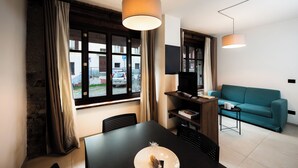 En moderne stue med en turkis sofa, et spisebord med stole, et fjernsyn monteret på væggen og et stort vindue med gardiner.