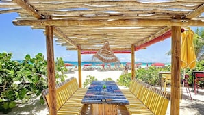 A beachside outdoor seating area with a table and chairs under a wooden pergola, overlooking the ocean.