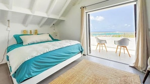 A bedroom with a bed, a view of the ocean, and a balcony with chairs.