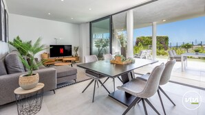 Ein modernes Wohnzimmer mit Flachbildfernseher, grauen Sofas, einem Esstisch mit Stühlen und einem großen Fenster mit Blick auf einen Garten.