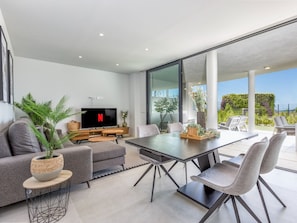 A modern living room with a flat-screen TV, grey sofas, a dining table with chairs, and a large window overlooking a garden. - Cameron | pool - garden - sea views (Fuengirola)