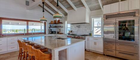 A spacious kitchen with a large island, white cabinetry, stainless steel appliances, and a view to the outside.