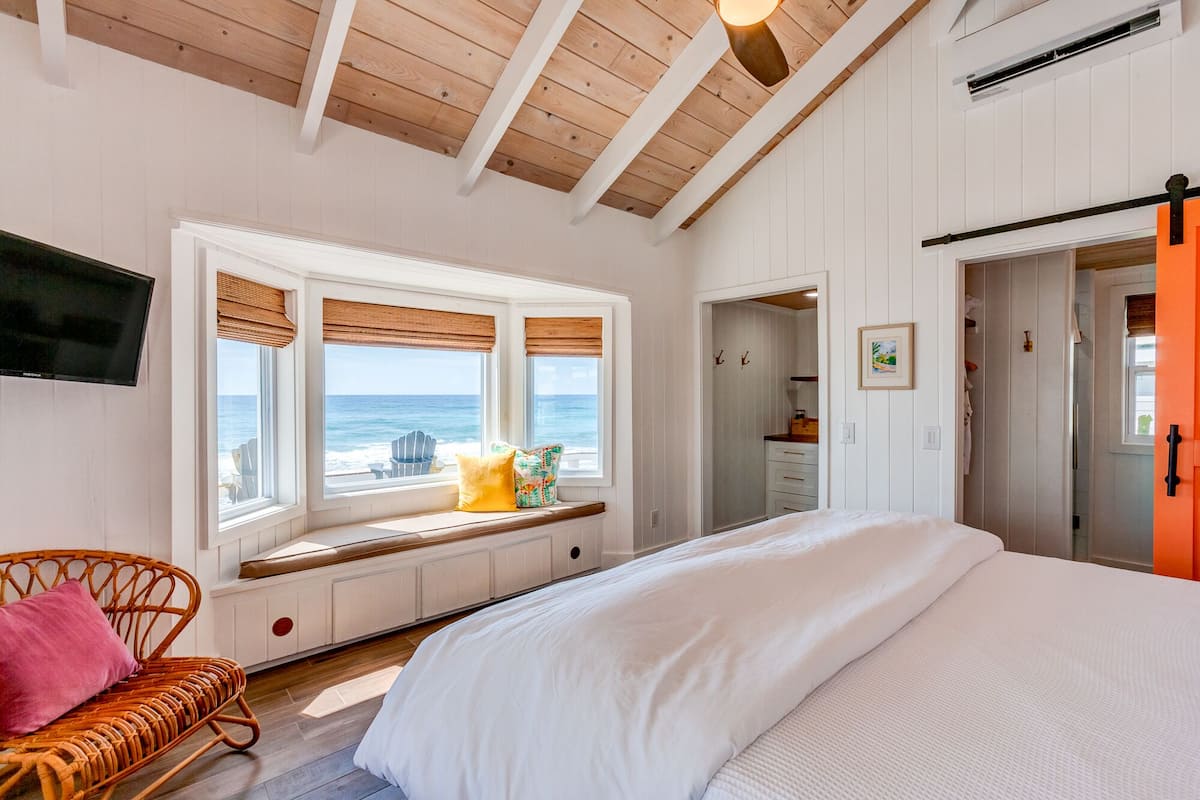 Un dormitorio con una cama grande, un banco junto a la ventana con vista al océano y una silla de mimbre.